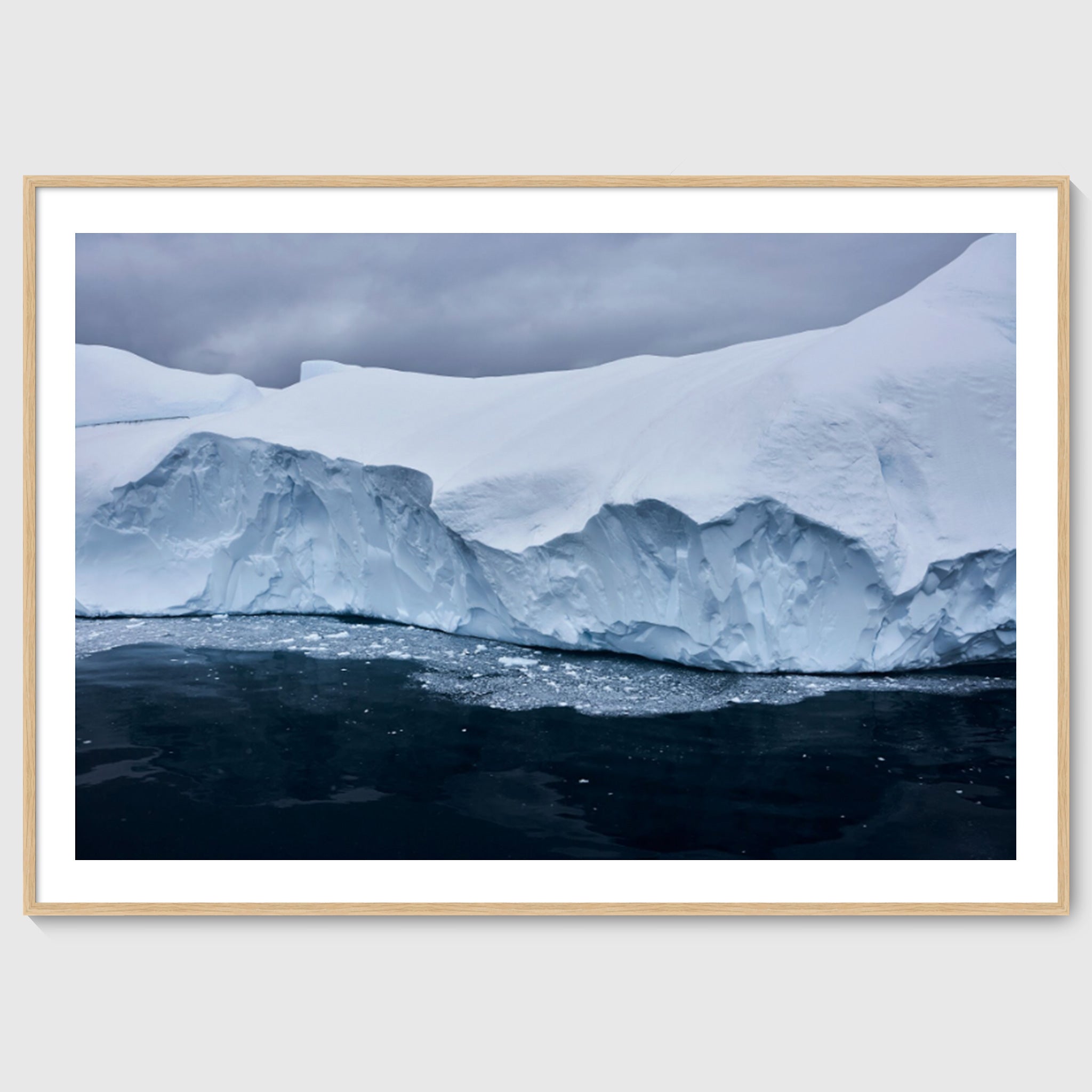Colour photograph of a glacier edge with fractured ice and dark water below, shown in an oak frame, Greenland