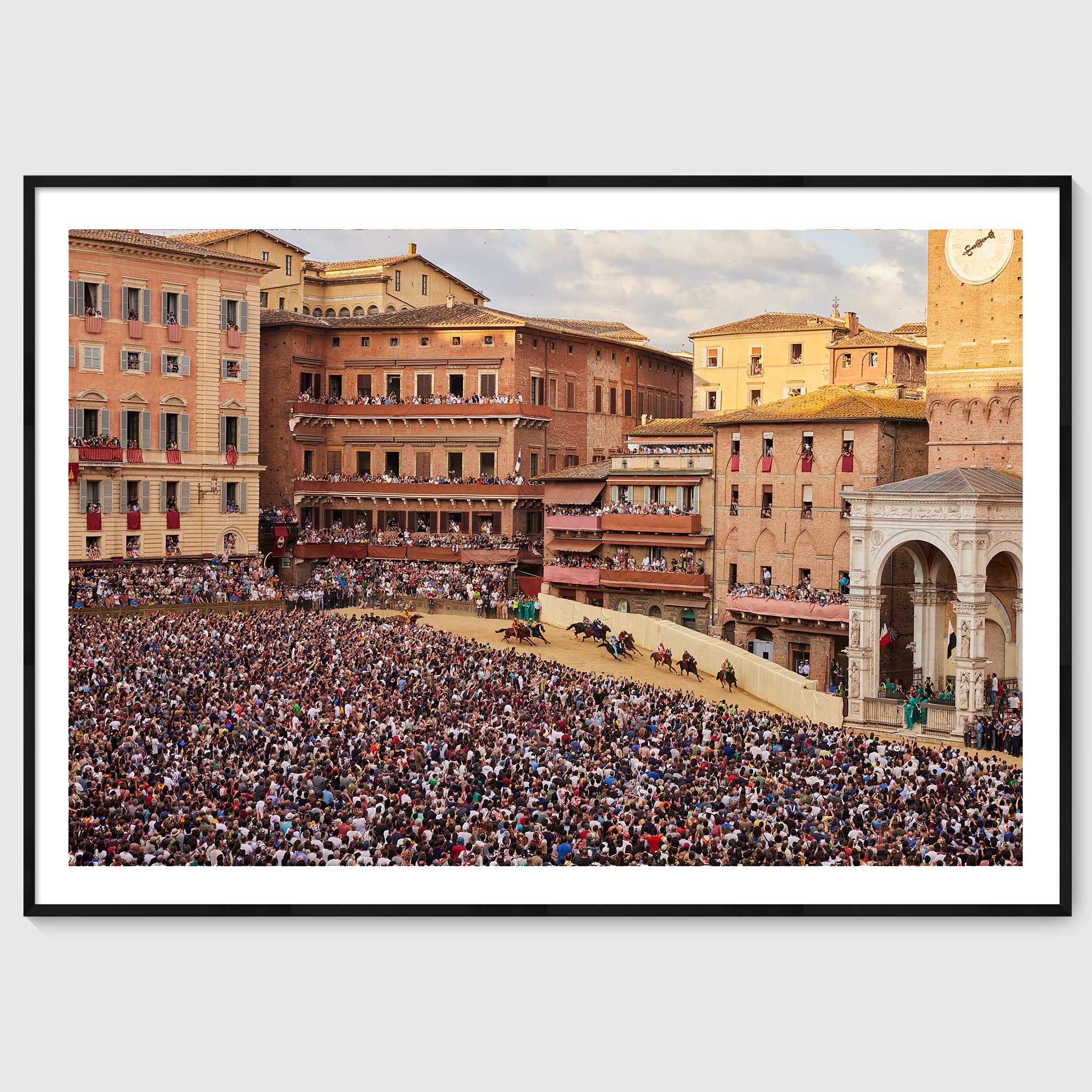 Colour photograph of jockeys on horseback rounding a tight bend in Piazza del Campo during the Palio di Siena, surrounded by densely packed crowds, shown in a black wooden frame