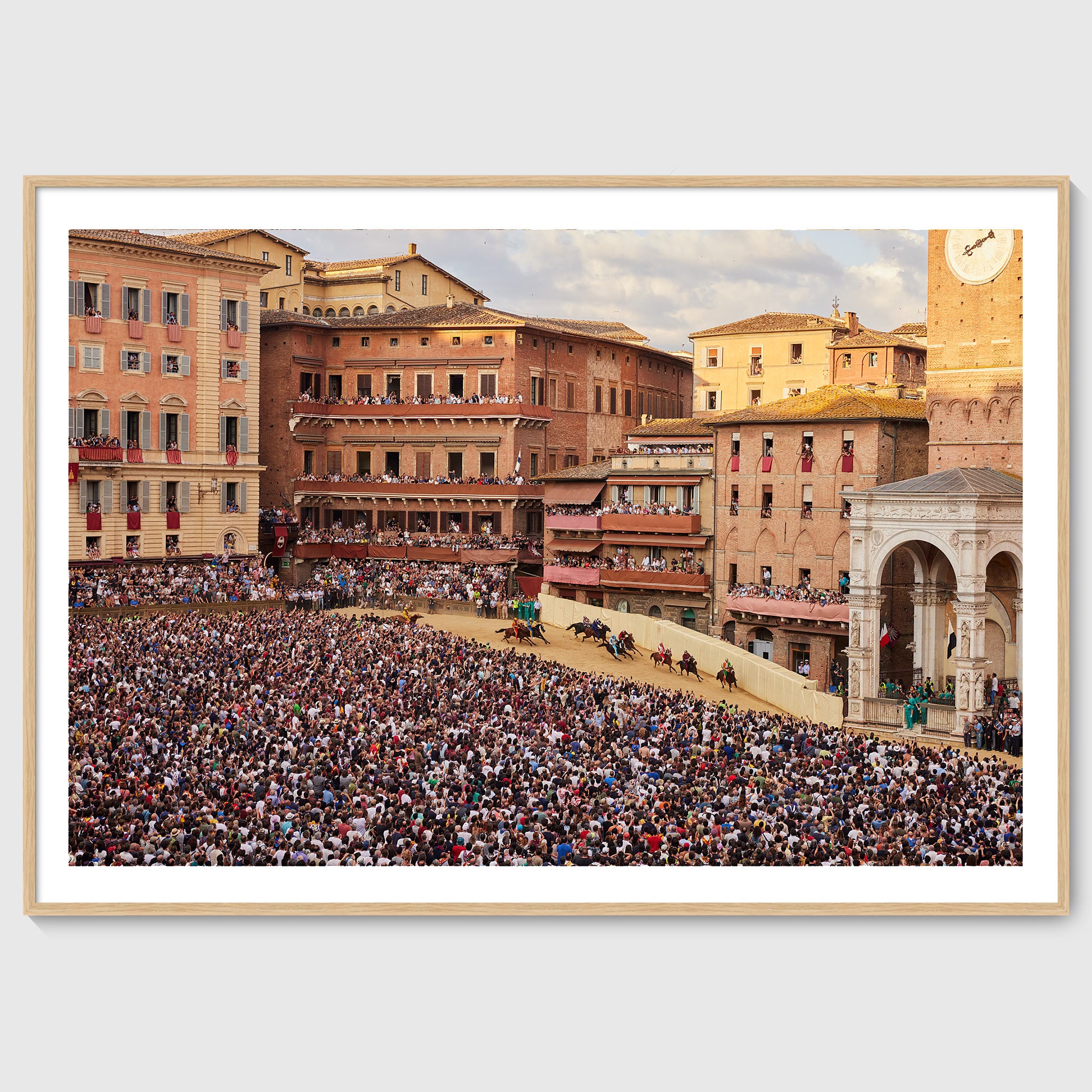 Colour photograph of jockeys on horseback rounding a tight bend in Piazza del Campo during the Palio di Siena, surrounded by densely packed crowds, shown in an oak wooden frame