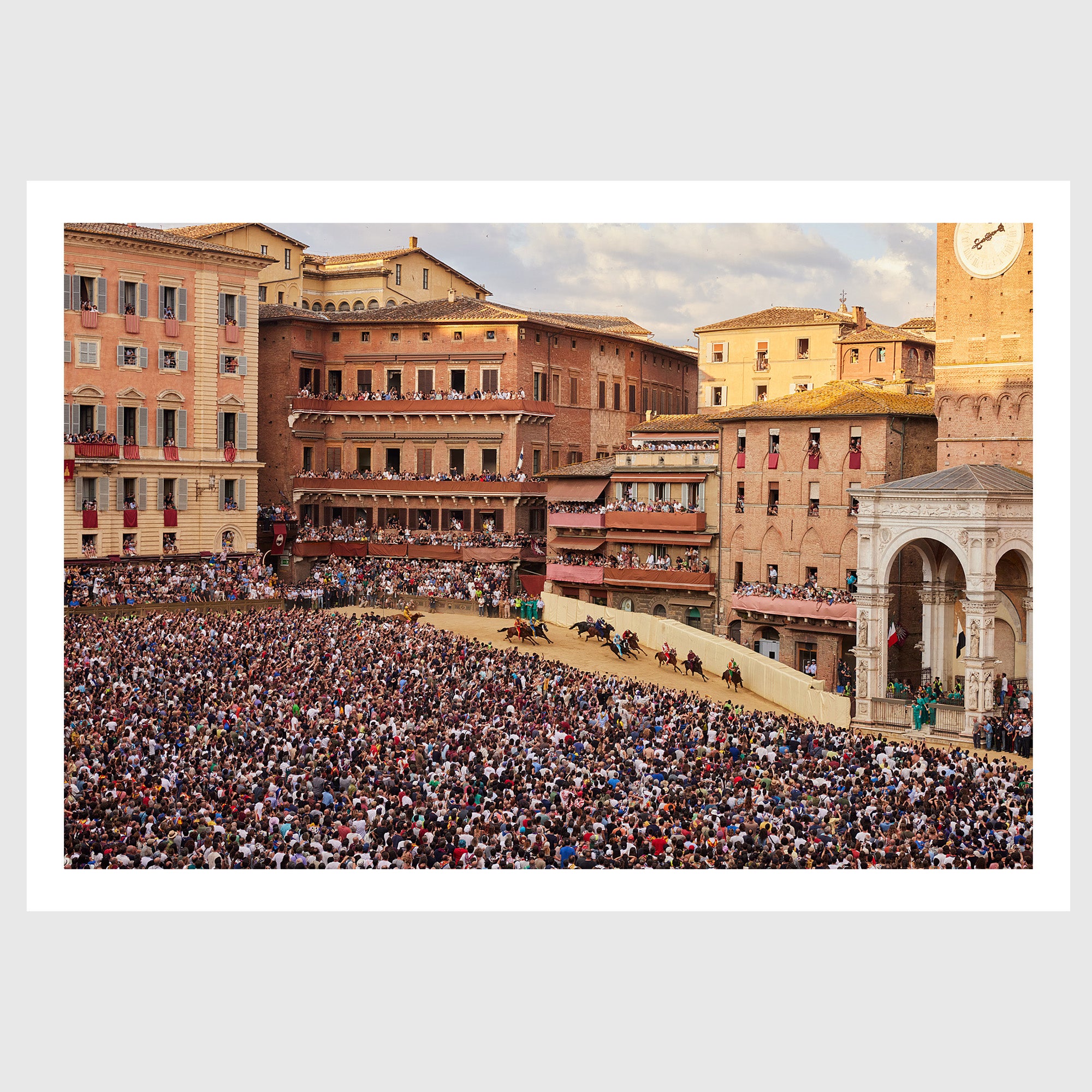 Colour photograph of jockeys on horseback rounding a tight bend in Piazza del Campo during the Palio di Siena, surrounded by densely packed crowds, sold as an unframed fine art print