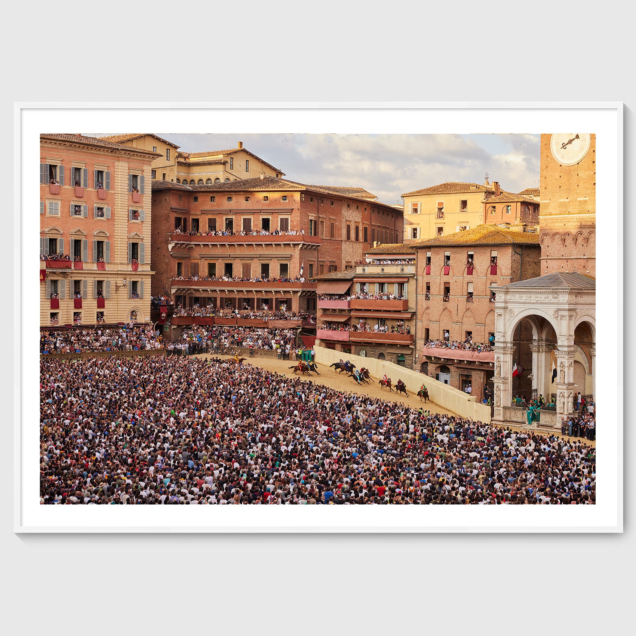 Colour photograph of jockeys on horseback rounding a tight bend in Piazza del Campo during the Palio di Siena, surrounded by densely packed crowds, shown in a white wooden frame
