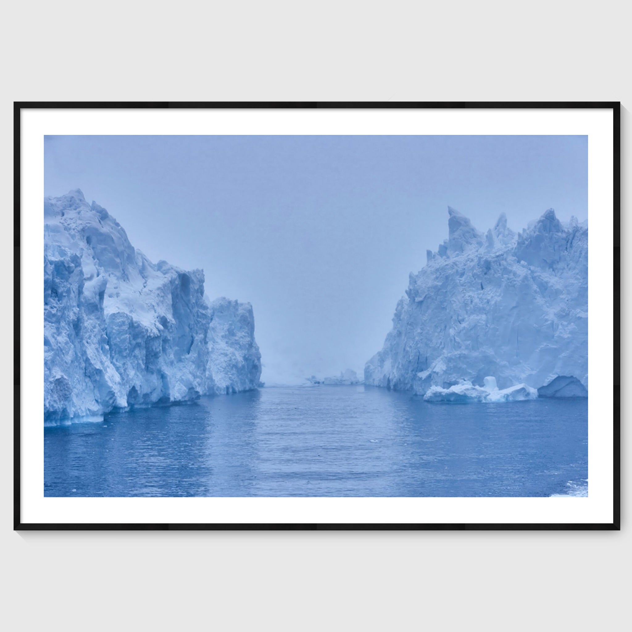 Colour photograph of a large iceberg rising from dark water beneath an overcast sky, shown in a black frame, Greenland