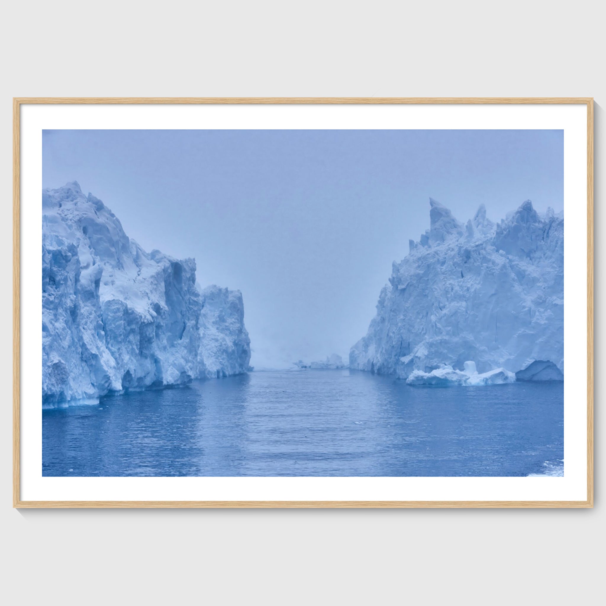 Colour photograph of a large iceberg rising from dark water beneath an overcast sky, shown in an oak frame, Greenland