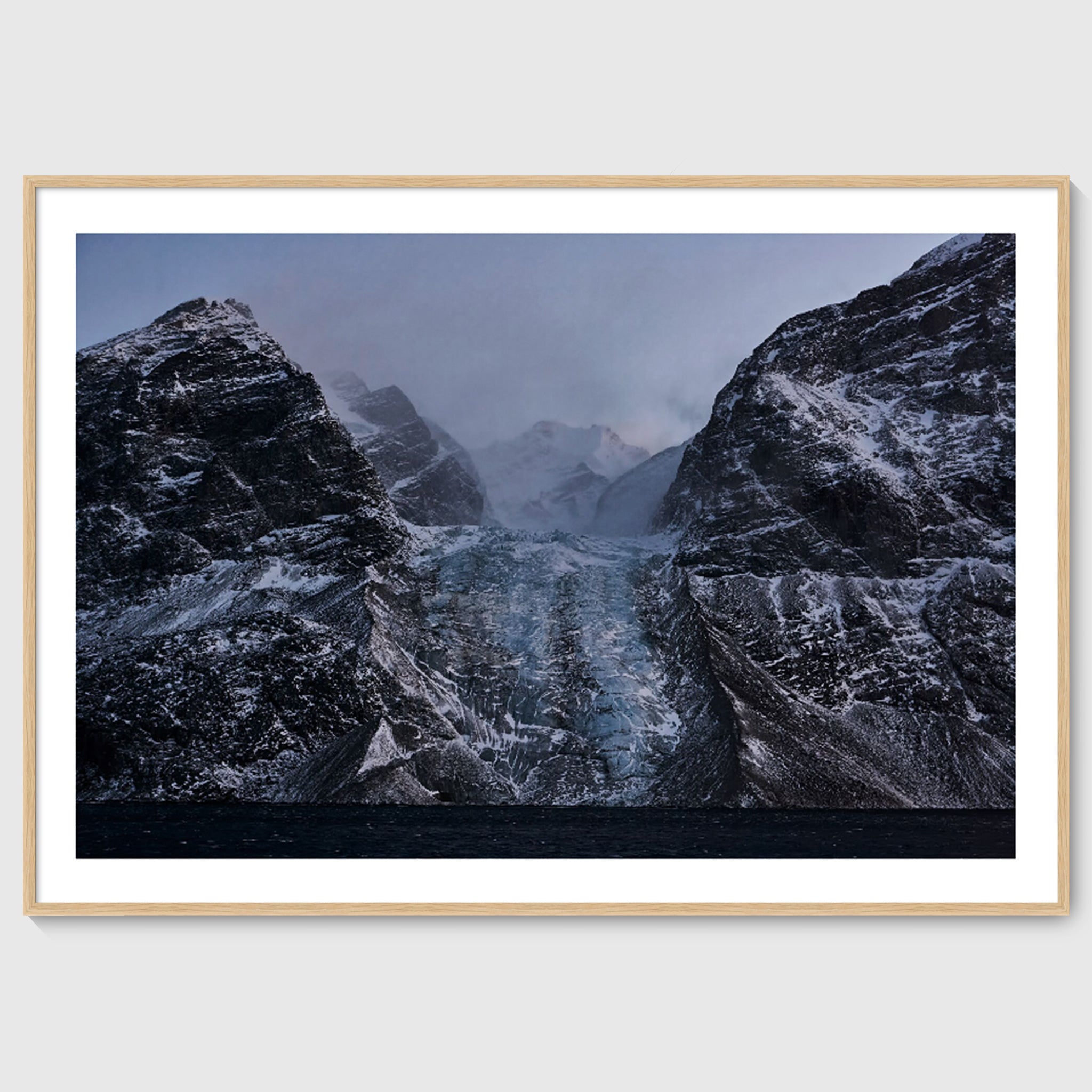 Colour photograph of a glacier descending between dark rock faces, shown in an oak frame, Greenland