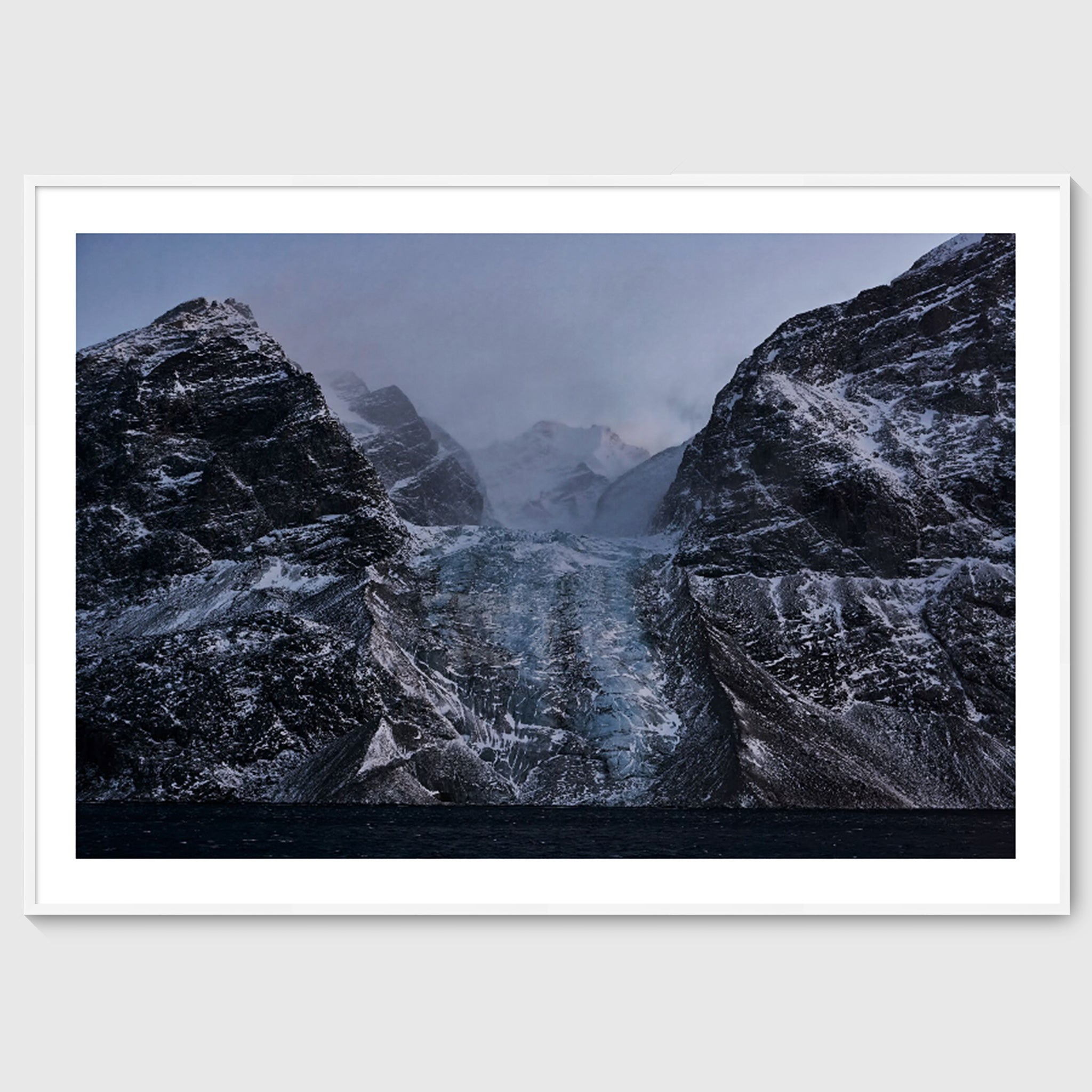 Colour photograph of a glacier descending between dark rock faces, shown in a white frame, Greenland