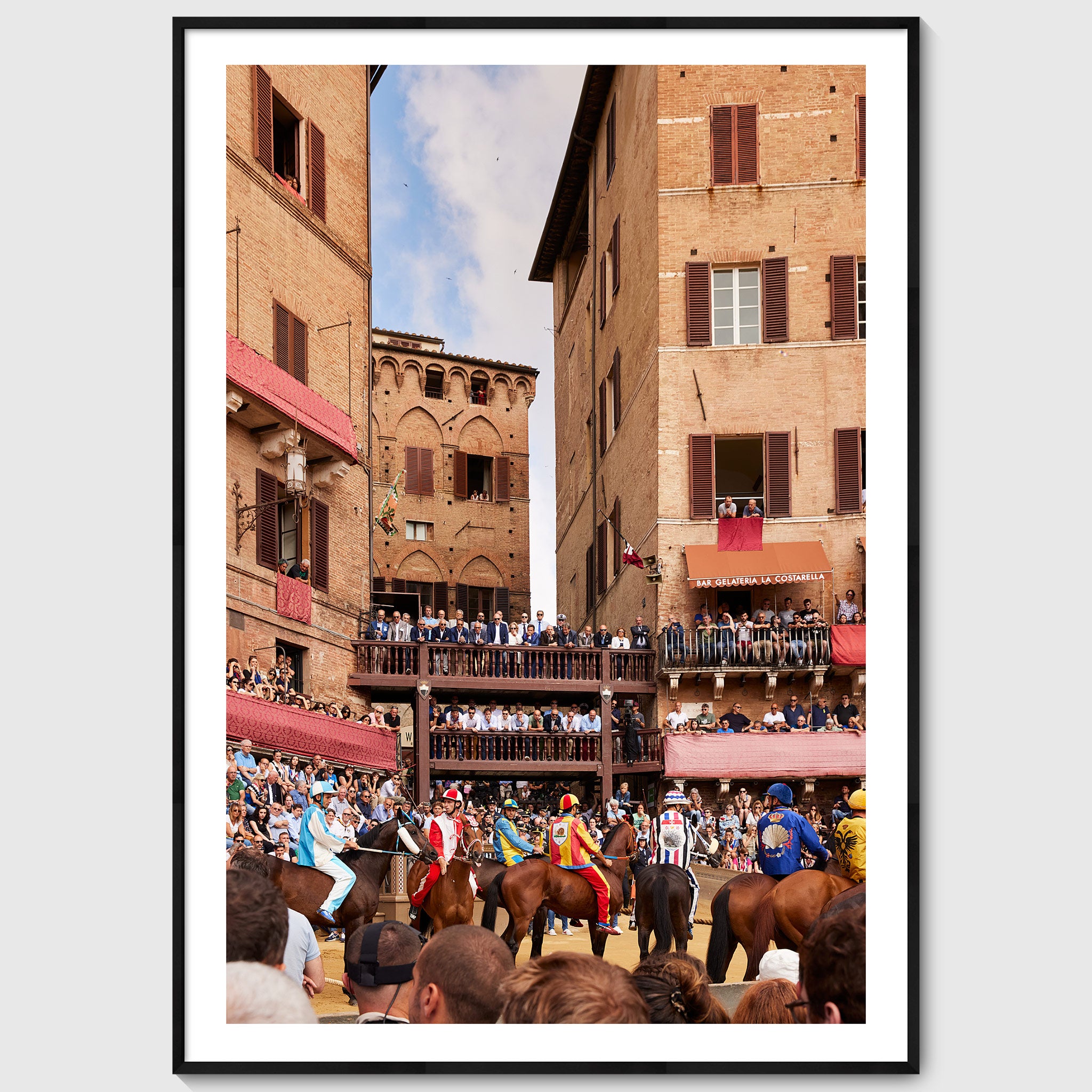 Colour photograph of jockeys on horseback lining up at the start of the Palio di Siena with crowds watching from balconies and the edges of Piazza del Campo, shown in a black wooden frame