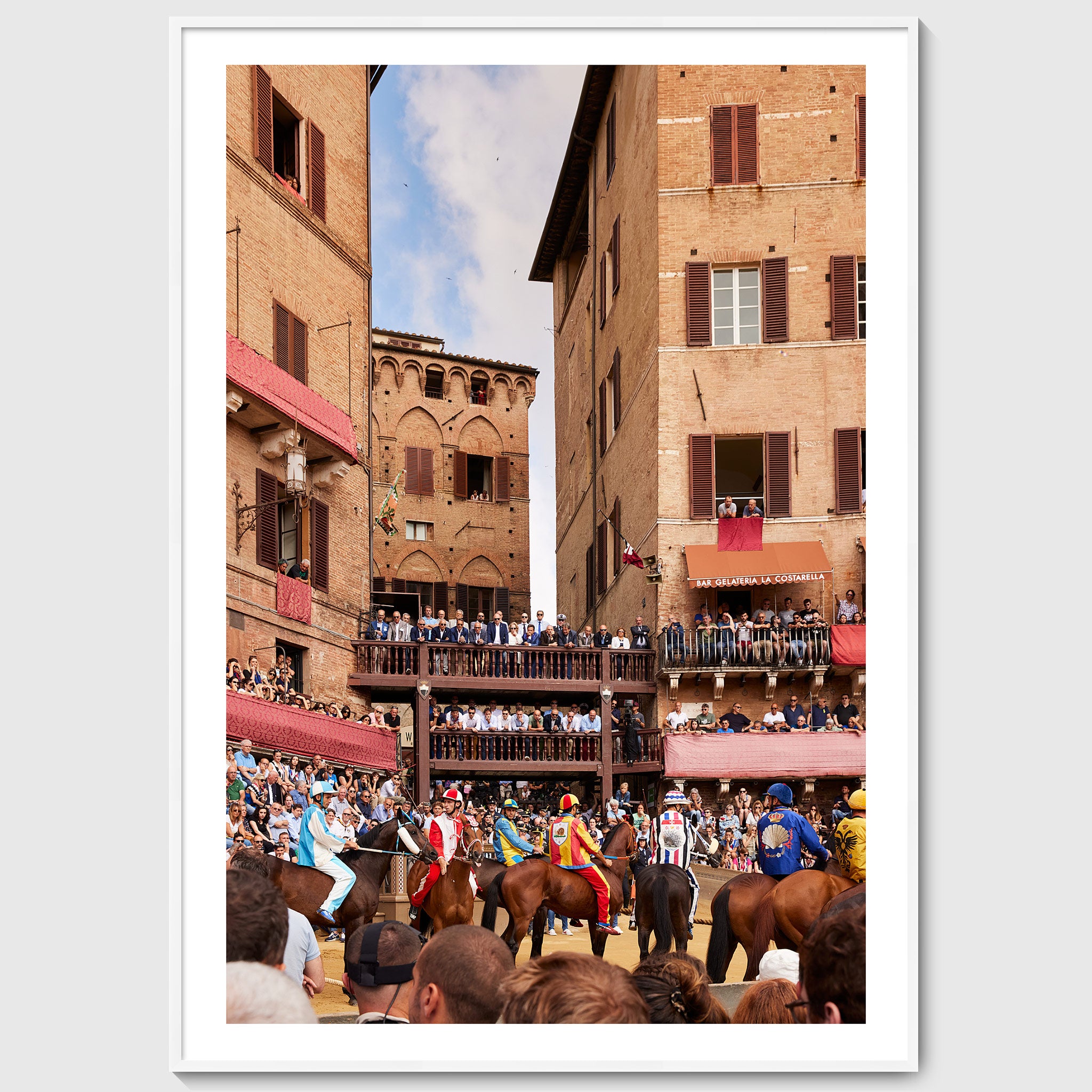 Colour photograph of jockeys on horseback lining up at the start of the Palio di Siena with crowds watching from balconies and the edges of Piazza del Campo, shown in a white wooden frame