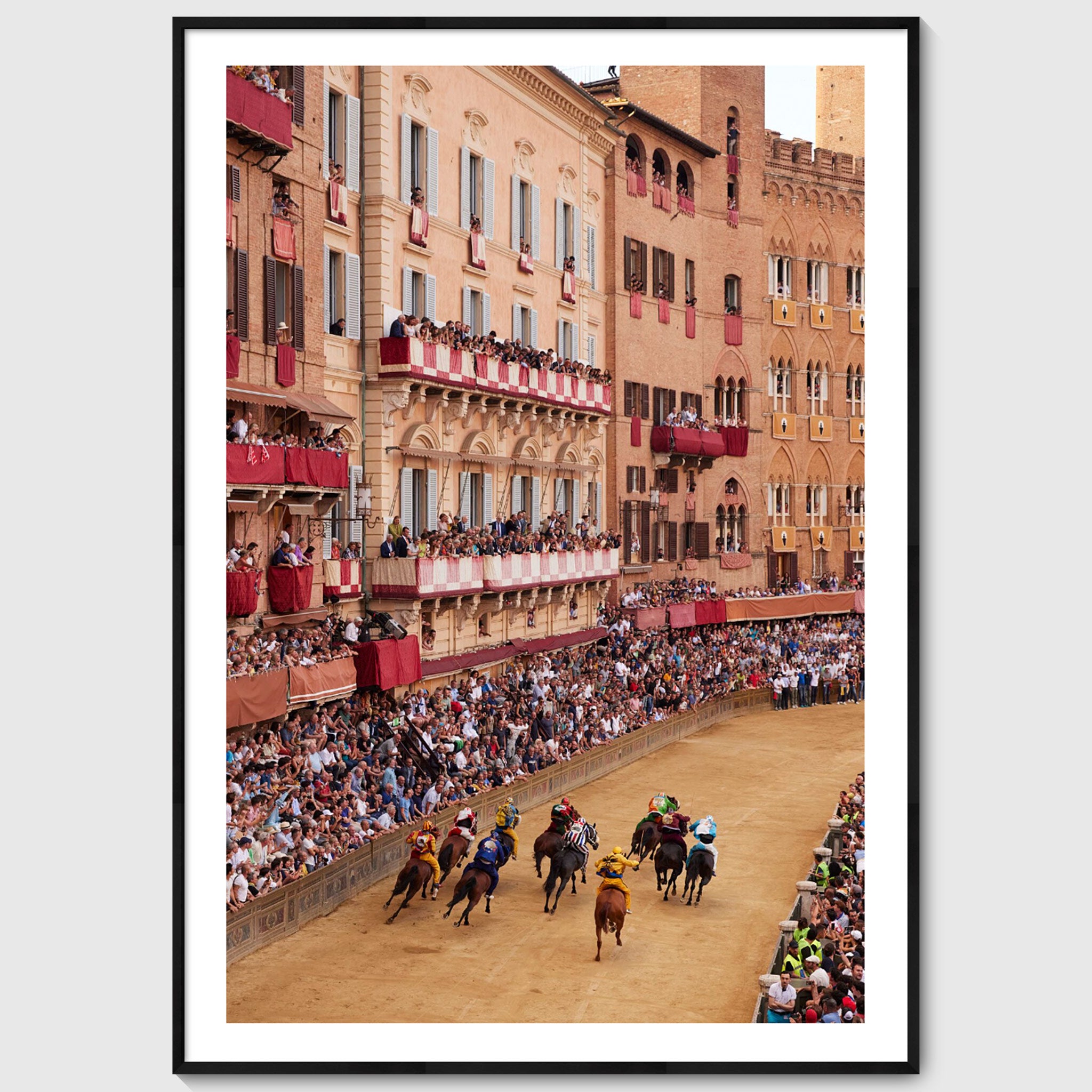 Colour photograph of Piazza del Campo filled with spectators as horses race past historic buildings during the Palio di Siena, shown in a black wooden frame