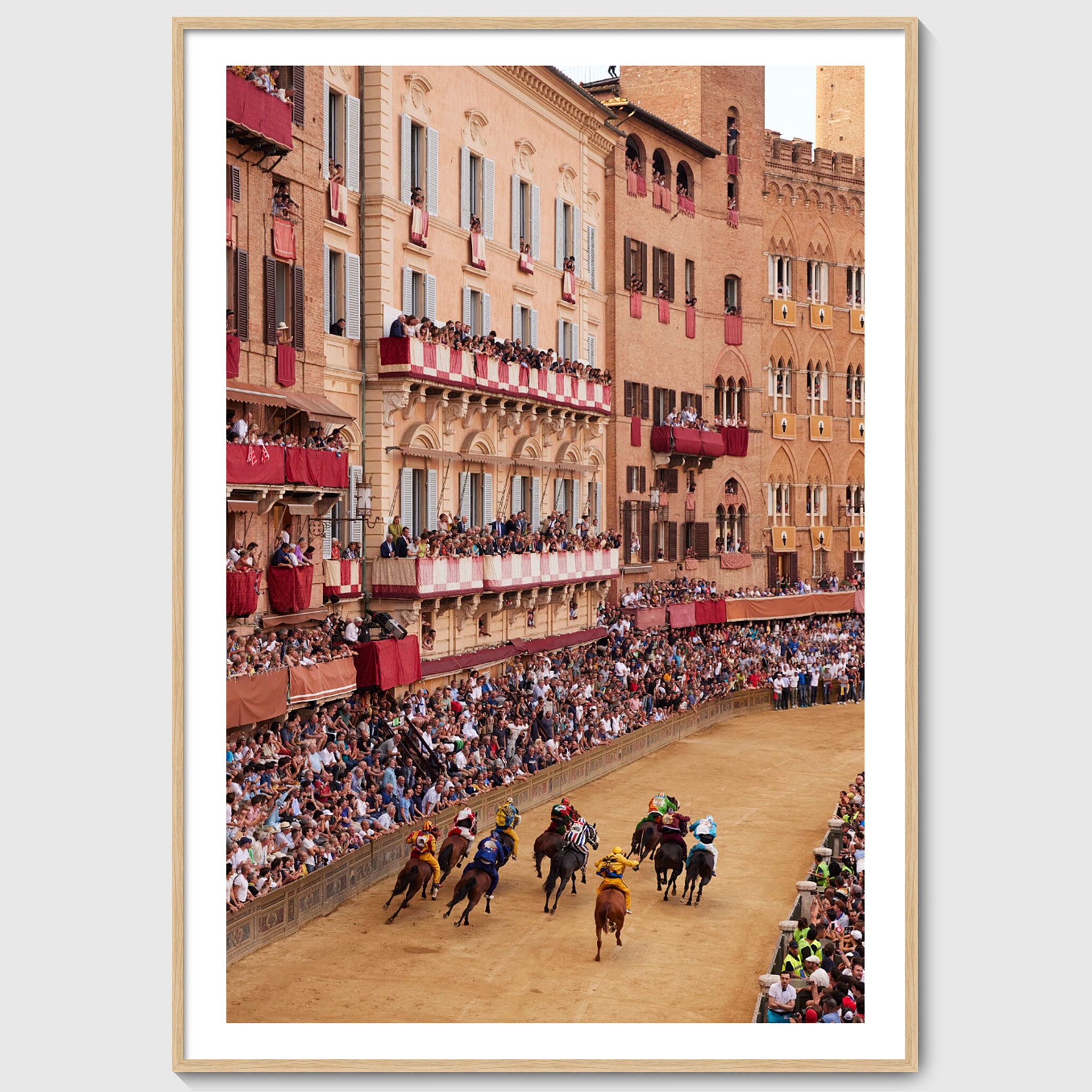 Colour photograph of Piazza del Campo filled with spectators as horses race past historic buildings during the Palio di Siena, shown in an oak wooden frame