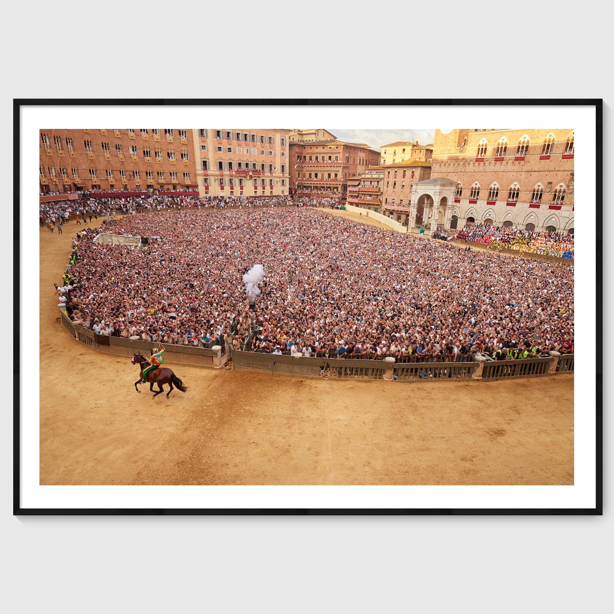 Colour photograph of a jockey on horseback riding past a dense crowd in Piazza del Campo during the Palio di Siena, shown in a black wooden frame