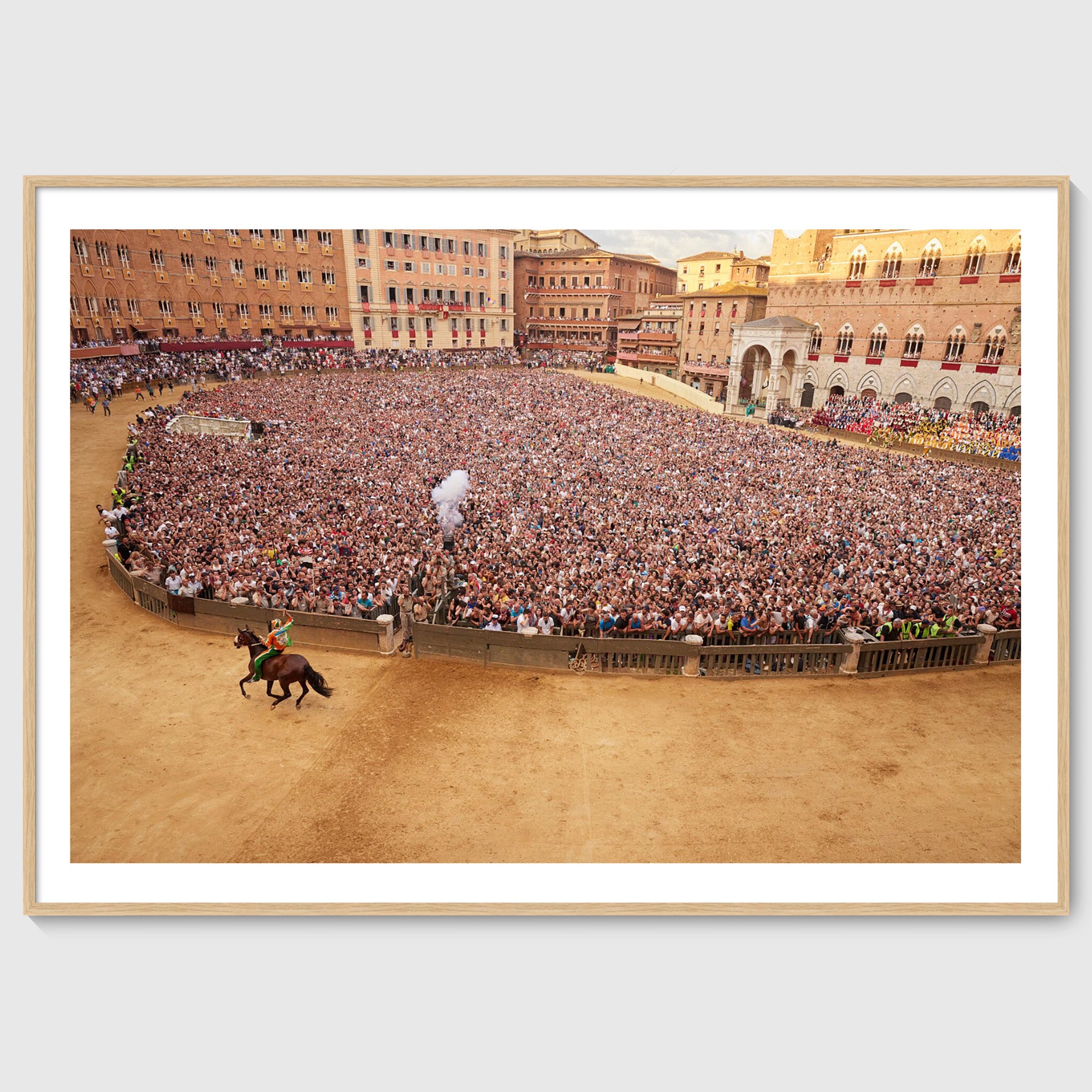 Colour photograph of a jockey on horseback riding past a dense crowd in Piazza del Campo during the Palio di Siena, shown in an oak wooden frame