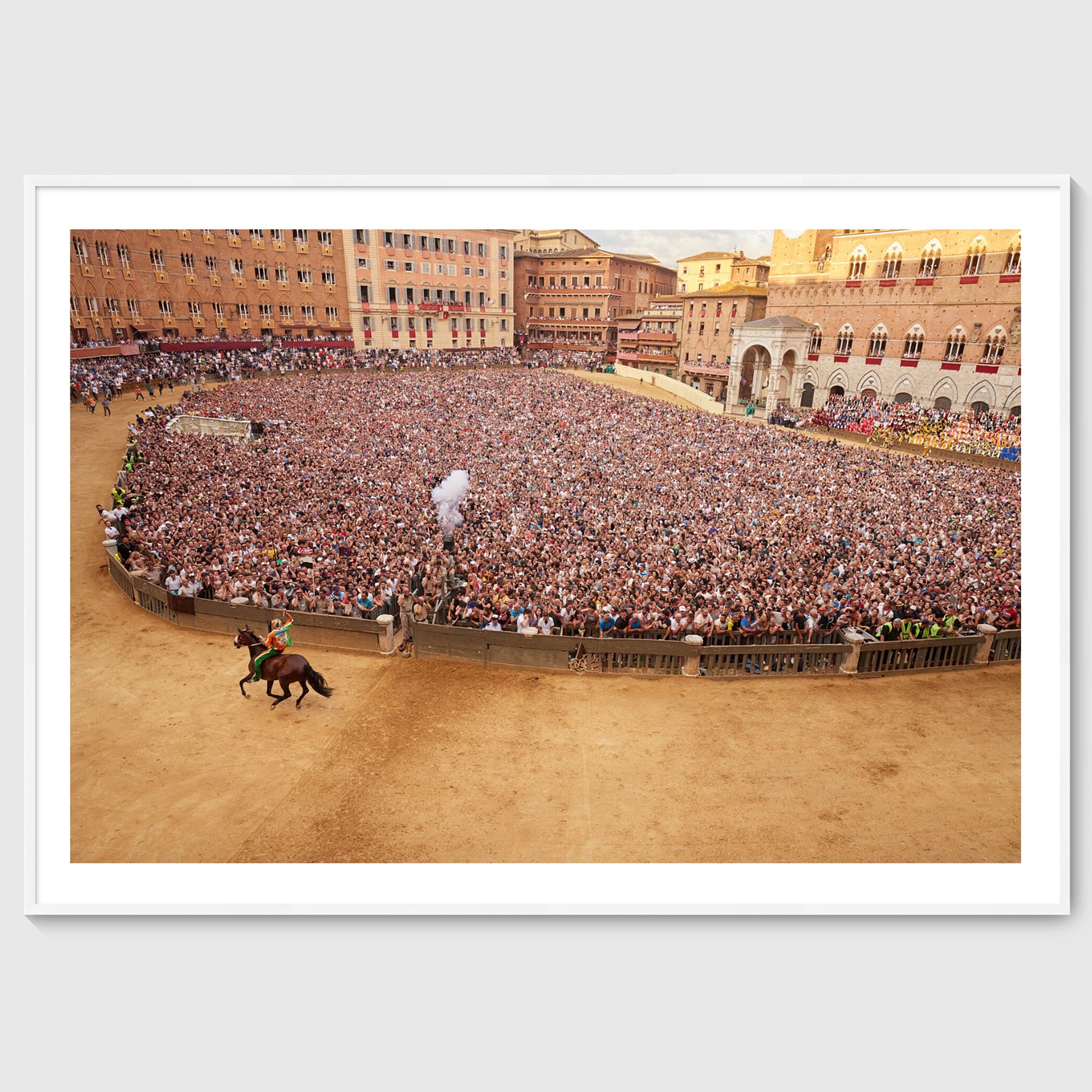 Colour photograph of a jockey on horseback riding past a dense crowd in Piazza del Campo during the Palio di Siena, shown in a white wooden frame